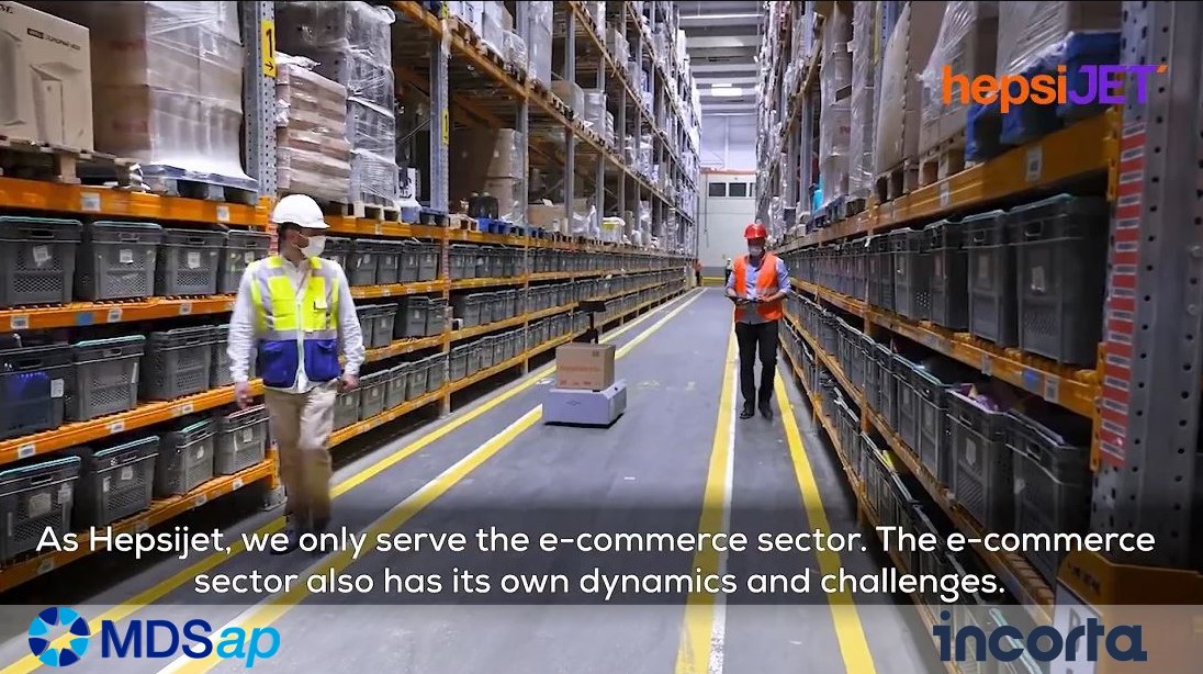 shows two men in an e-commerce warehouse with a robot in the middle distance