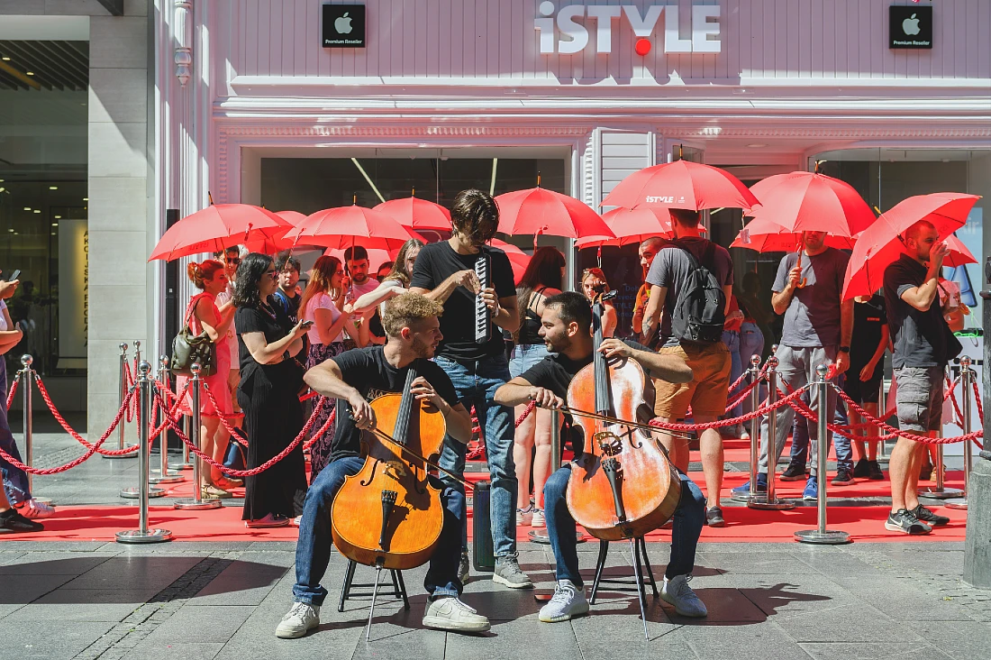 three musicians entertain a queue of people sheltering from the sun under red parasols