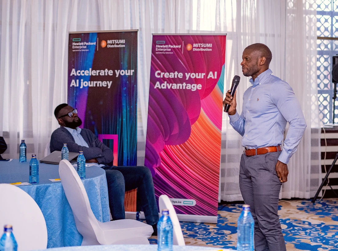 a man speaks into a mic at a technology event while a seated man looks onwards.