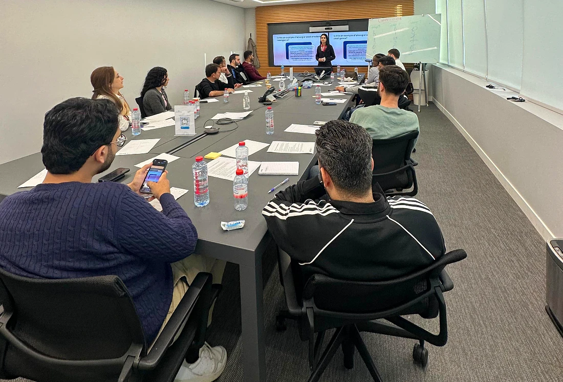 a large group of people are sitting around a boardroom table watching a trainer in from of a presentation screen.